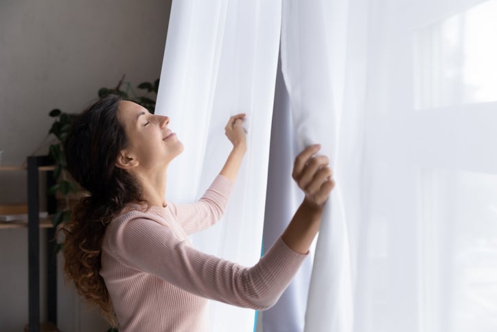 A woman stands in front of an open window, enjoying the incoming breeze.